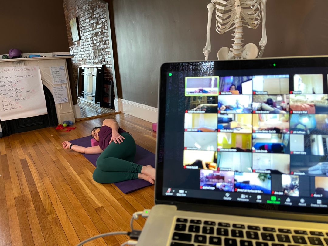 In the background Nancy Bellantoni on the floor demonstrating how to use therapy balls. Part of a skeleton is visible over a computer with students in Zoom.